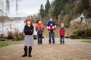 Four children standing on a brick path near a lake with evergreen trees in the background.