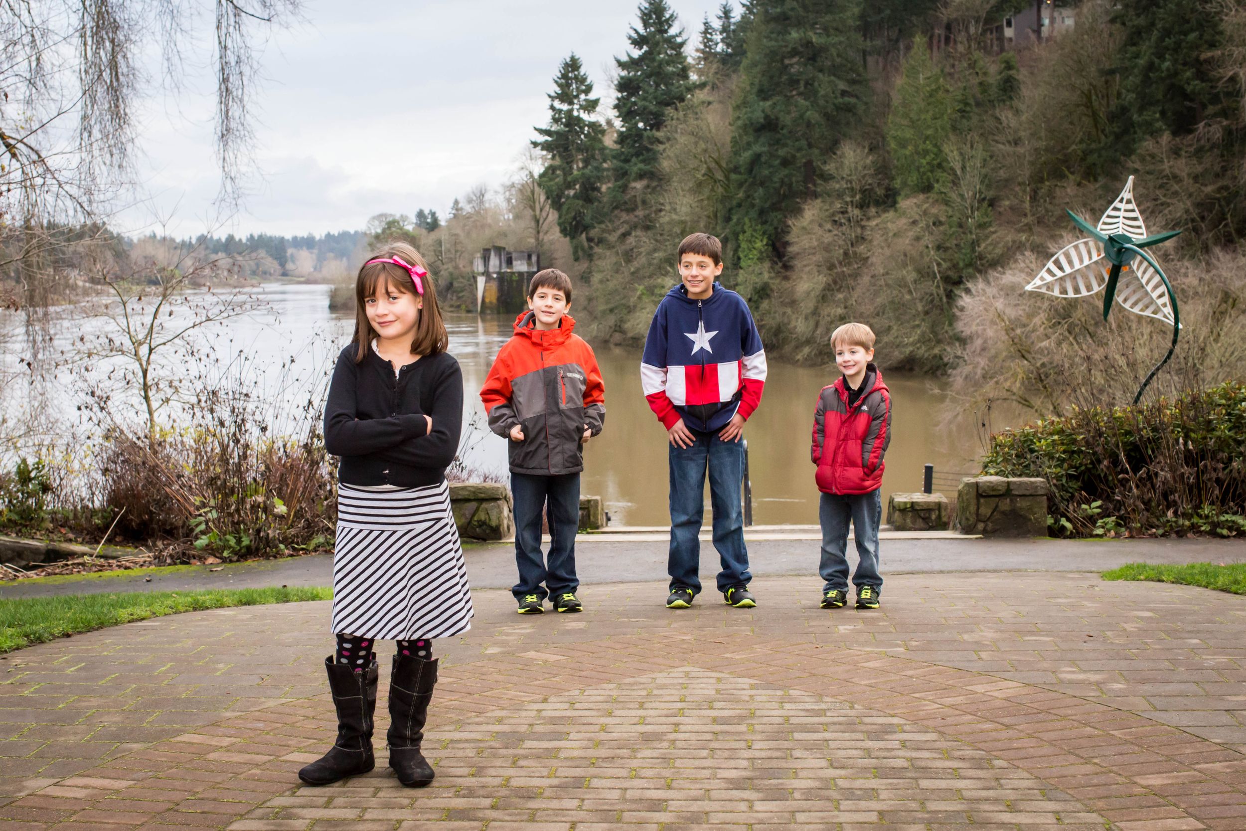 Four children standing on a brick path near a lake with evergreen trees in the background.