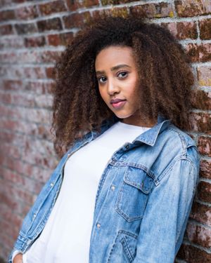 Stylish woman in denim jacket with natural curly hair posing against brick wall background.