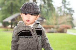 A toddler wearing a gray knit cap and jacket stands in a grassy yard.