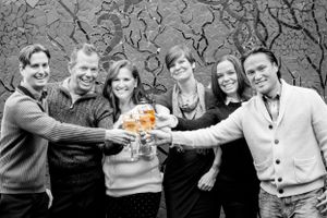 A black and white photo of friends toasting drinks together outdoors with one orange-colored glass.
