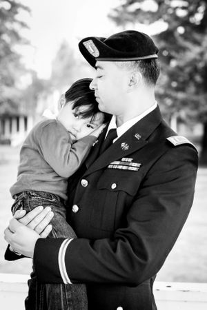 Military service member in dress uniform holds a young child in black and white portrait.