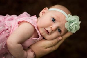 A baby in a pink dress with a mint green headband lying on soft hands.