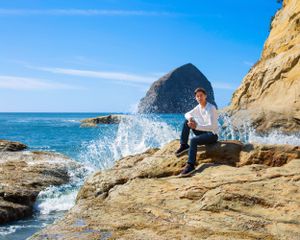 Person sitting on coastal rocks with dramatic ocean waves splashing and mountain peak in background.