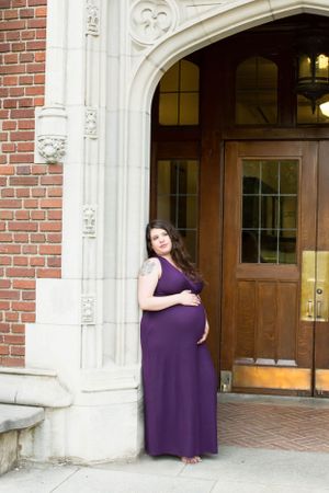 Expectant mother in purple maxi dress poses elegantly by brick building entrance.