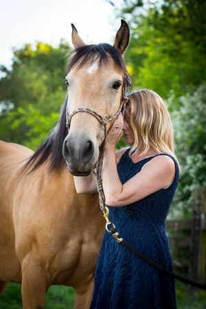 A woman in a navy dress whispers to a brown horse wearing a bridle outdoors.