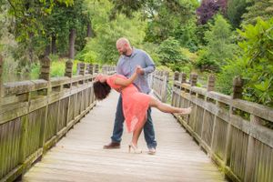 Couple dancing on wooden bridge with romantic dip pose surrounded by lush greenery.