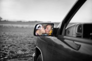 Side mirror of a car showing a young child looking out the window onto a field.