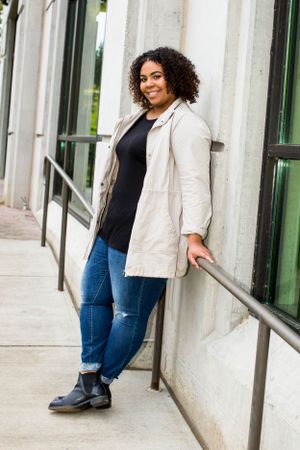 Casual portrait of woman in beige jacket and jeans leaning against building wall.