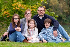 Family of five smiling outdoors on grass with autumn trees in background.