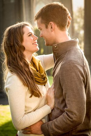 A happy young couple embraces outdoors in warm autumn light, smiling at each other romantically.