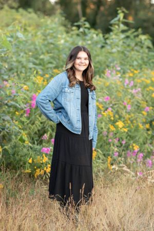 Woman in denim jacket and black maxi dress poses in wildflower meadow at golden hour.