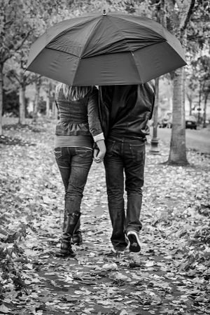 A couple walking together under a black umbrella on a path covered in fallen autumn leaves.