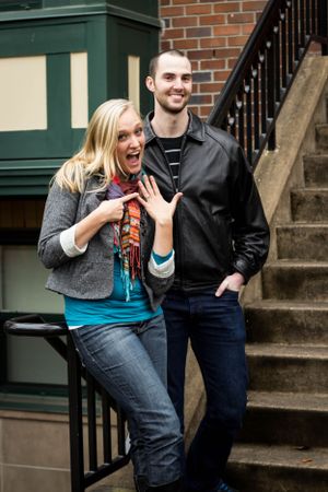 Excited couple on urban steps showing off engagement ring in casual attire.