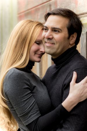 Romantic couple in dark sweaters embrace against wall in intimate portrait.