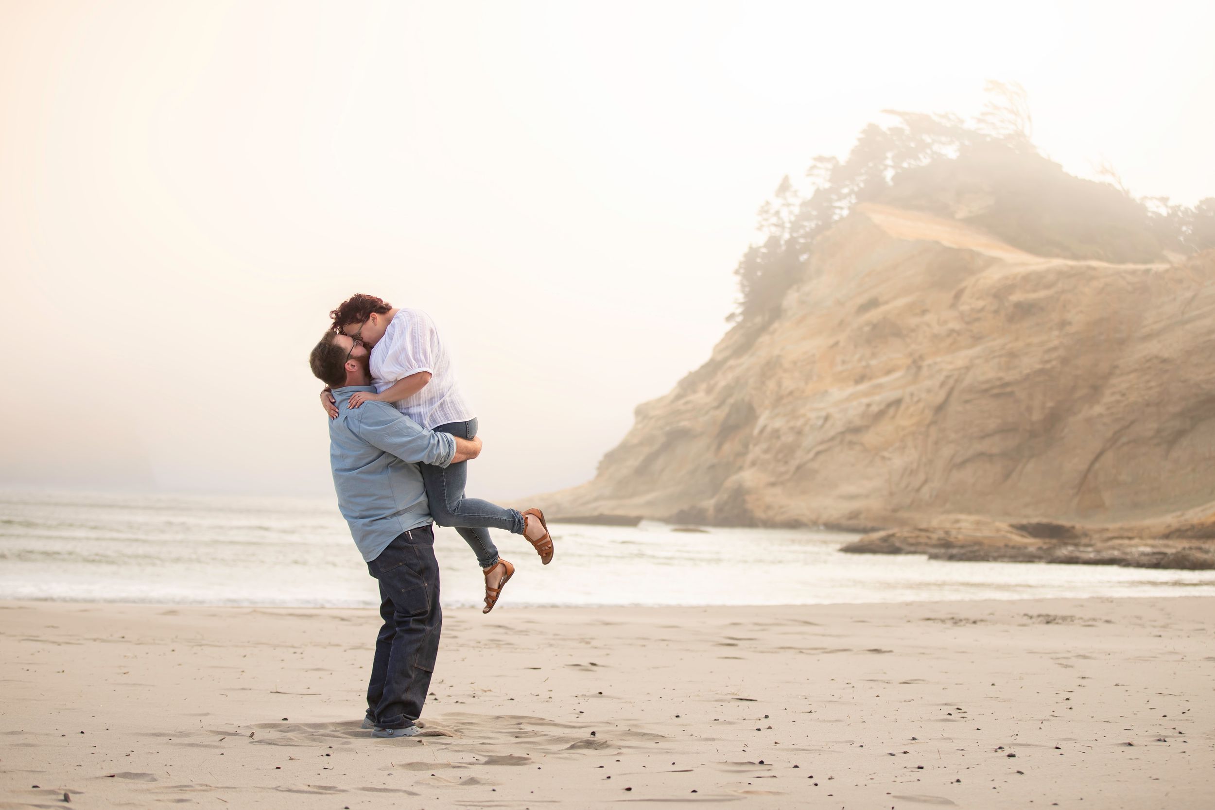 A couple embraces on a misty beach with dramatic cliffs and mountains in the background during sunset.