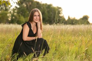 Someone in a black dress sits in a golden sunlit meadow during sunset with tall grass surrounding them.