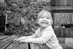 Black and white close-up of smiling child leaning on rustic wooden table near stone wall.