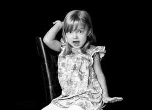 Black and white portrait of small child making expressive face while sitting on chair.