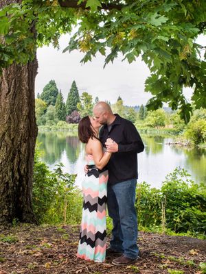 Couple sharing a romantic kiss under a large tree by a serene lake in a lush green park setting.