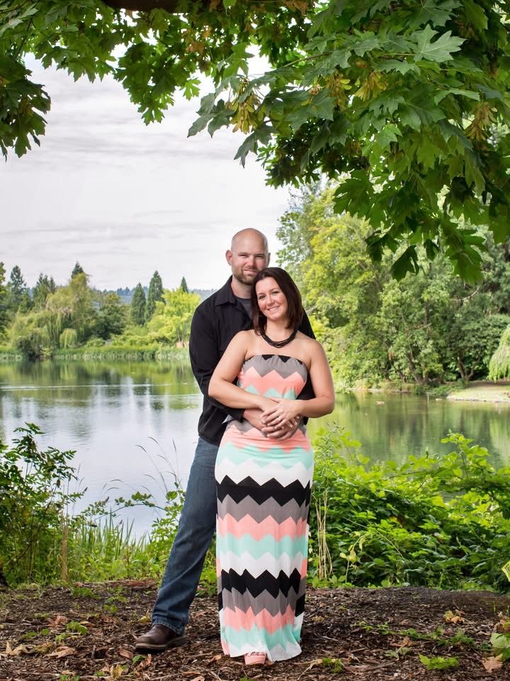 Couple posing romantically by a serene river surrounded by lush green trees and foliage in an outdoor portrait.