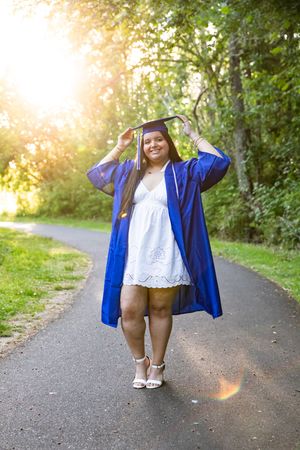 Young woman in blue graduation cap and gown smiling on a tree-lined path in golden sunlight.