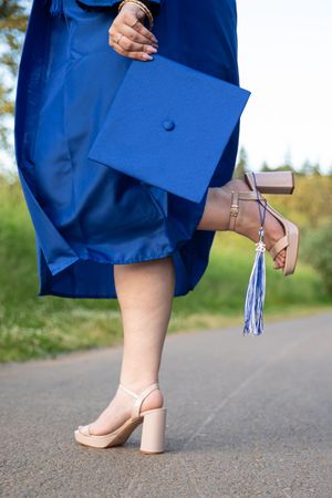 Graduate in blue cap and gown holding mortarboard, wearing nude block heel sandals on paved path outdoors.