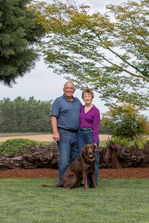 A couple poses outdoors on a lawn with their chocolate Labrador dog, with trees and landscaping in the background.
