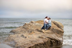 A couple embraces on a large coastal rock formation with ocean waves crashing in the background.