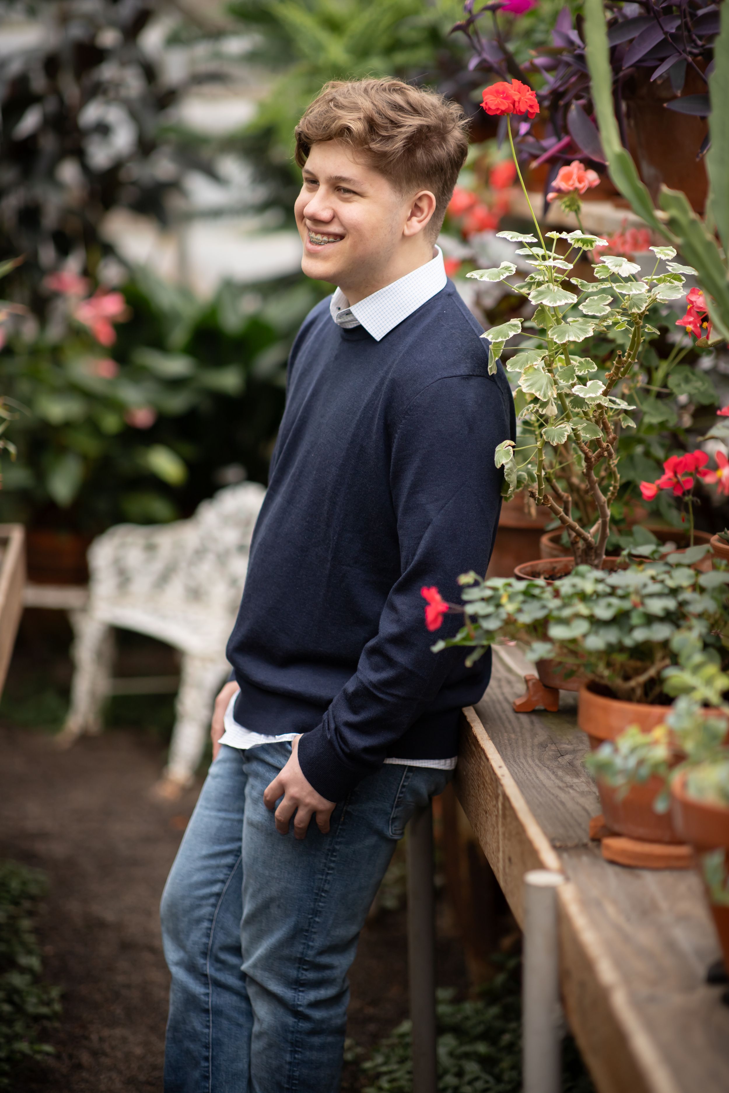 A young student in navy sweater and gray pants stands in a garden with potted flowers and plants.