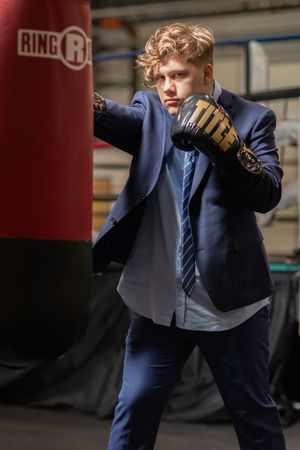 Professional boxer in navy suit throws practice punches at a boxing gym with red punching bags visible in background.