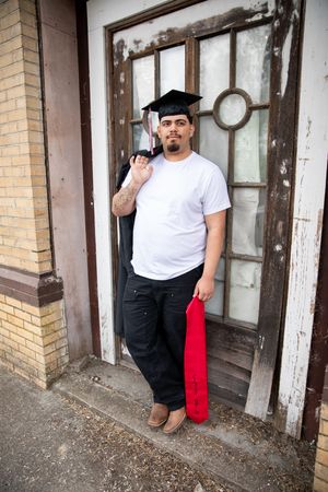 Young man in graduation cap holding red diploma cover, posing proudly in front of rustic building door.
