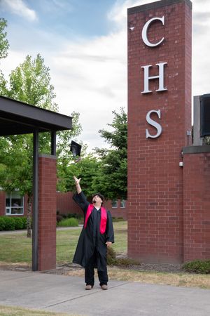Graduate in black and red gown tosses cap in air outside CHS brick school entrance on cloudy day.