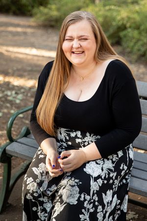 Smiling young woman with long blonde hair sitting on a park bench wearing a black top and floral skirt.