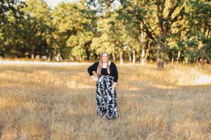 Woman in black top and floral maxi skirt standing in golden meadow with oak trees in background.