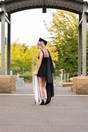 A graduate in black cap and gown poses under an archway, holding a diploma, with green trees in the background.