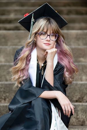Young woman in graduation cap and gown with colorful hair and glasses sits thoughtfully on stone steps.