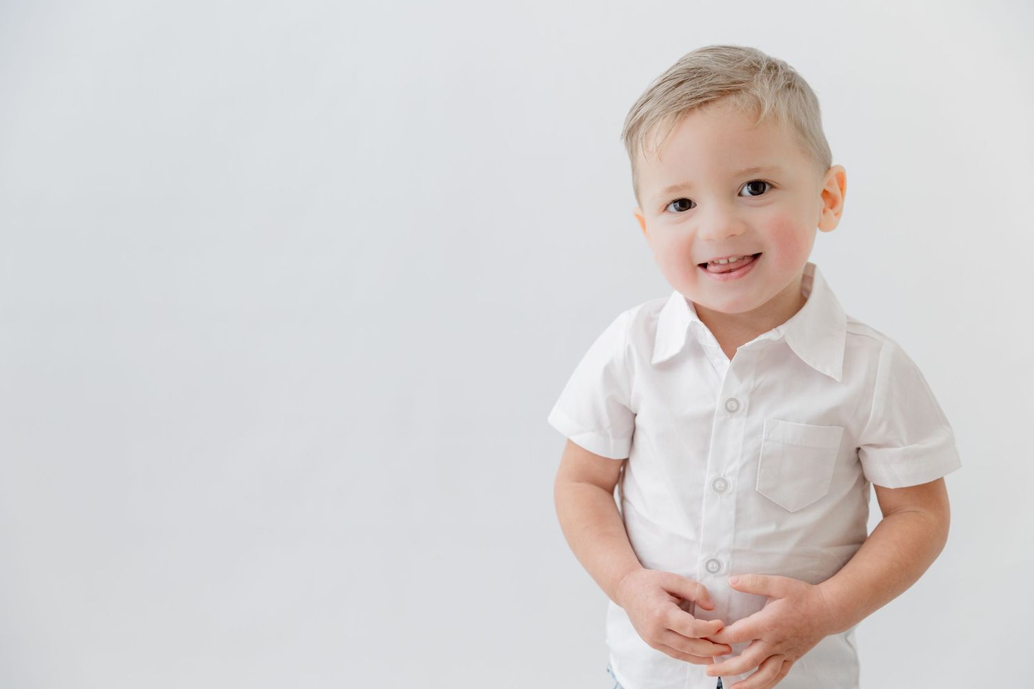 Smiling toddler boy in white button-up shirt posing against a white background.