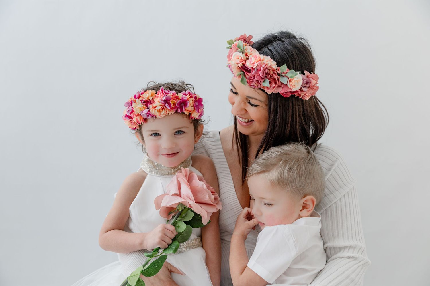 A smiling mother and two young children wearing pink floral crowns pose together against a white background.