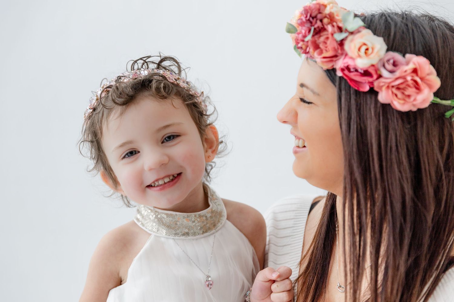 A mother wearing a floral crown holds her smiling toddler daughter, both dressed in white, against a light background.