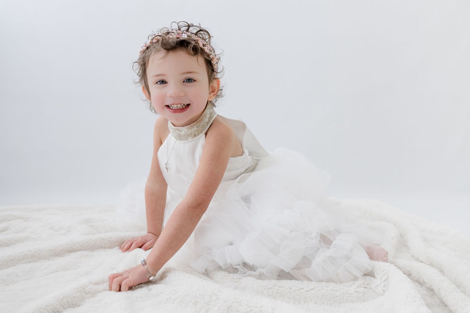 A smiling toddler girl in a white tulle dress and floral headband poses on a white fluffy blanket.