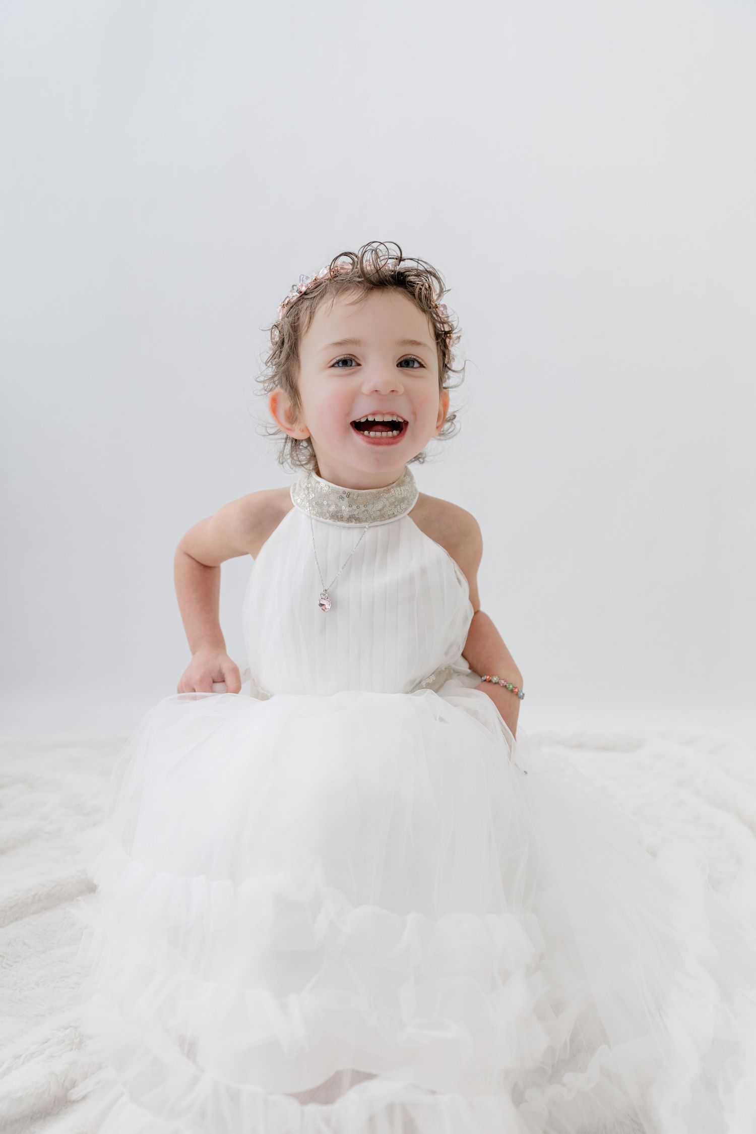 Smiling toddler girl in a white halter dress with curly hair, sitting on a white background.