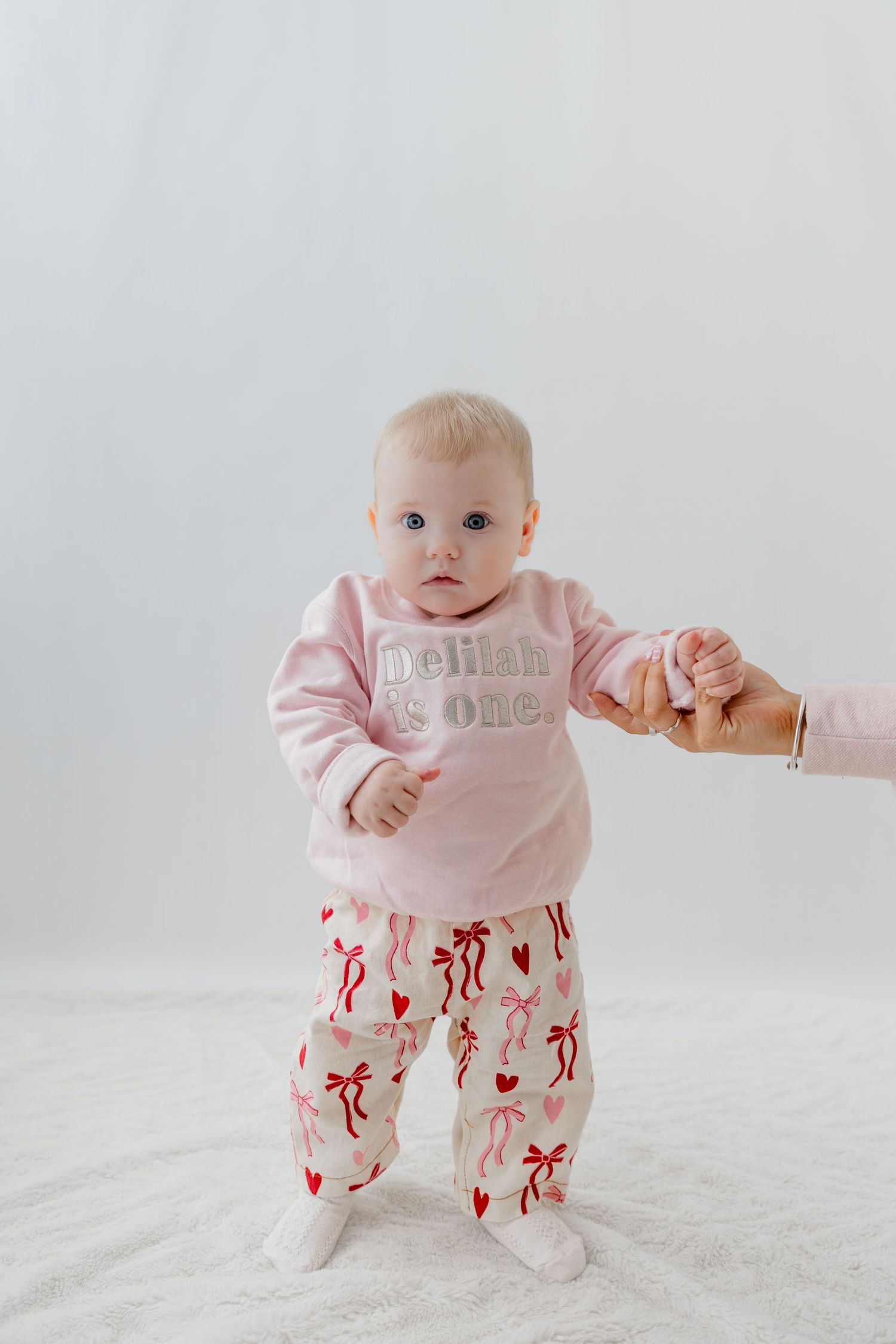 Baby girl in pink "Delilah is One" sweatshirt and bow-print pants standing with adult hand support.