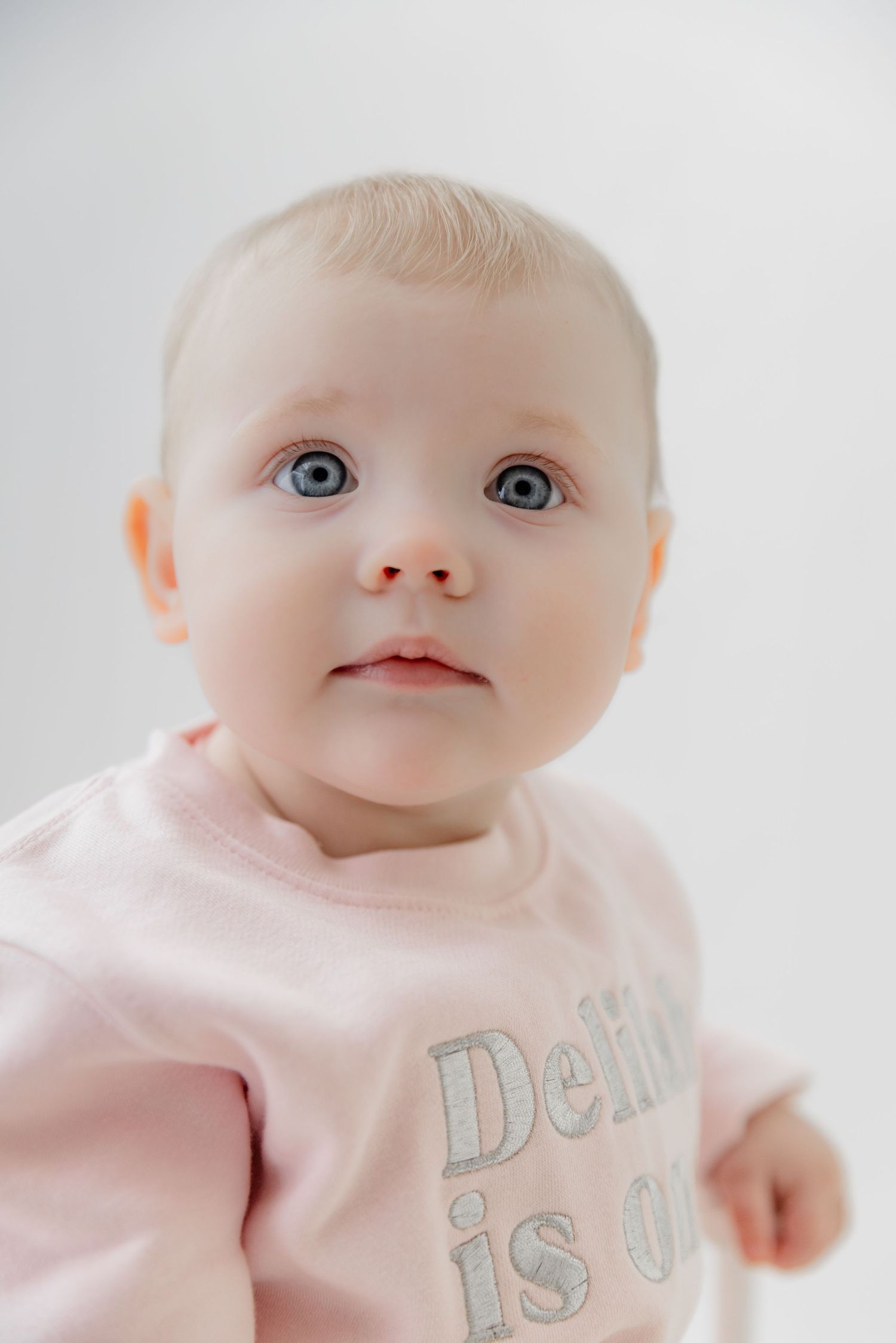 A cute baby with blue eyes wearing a pink shirt sits against a white background, looking up with curiosity.