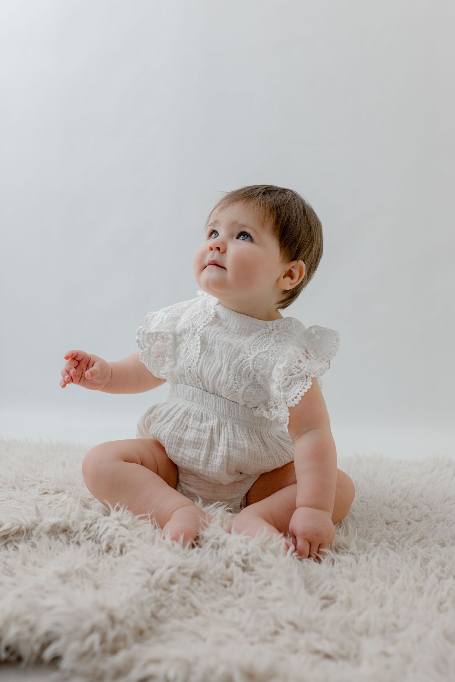 A baby girl in a white lace romper sits on a fluffy cream rug, gazing upward in a bright, airy setting.