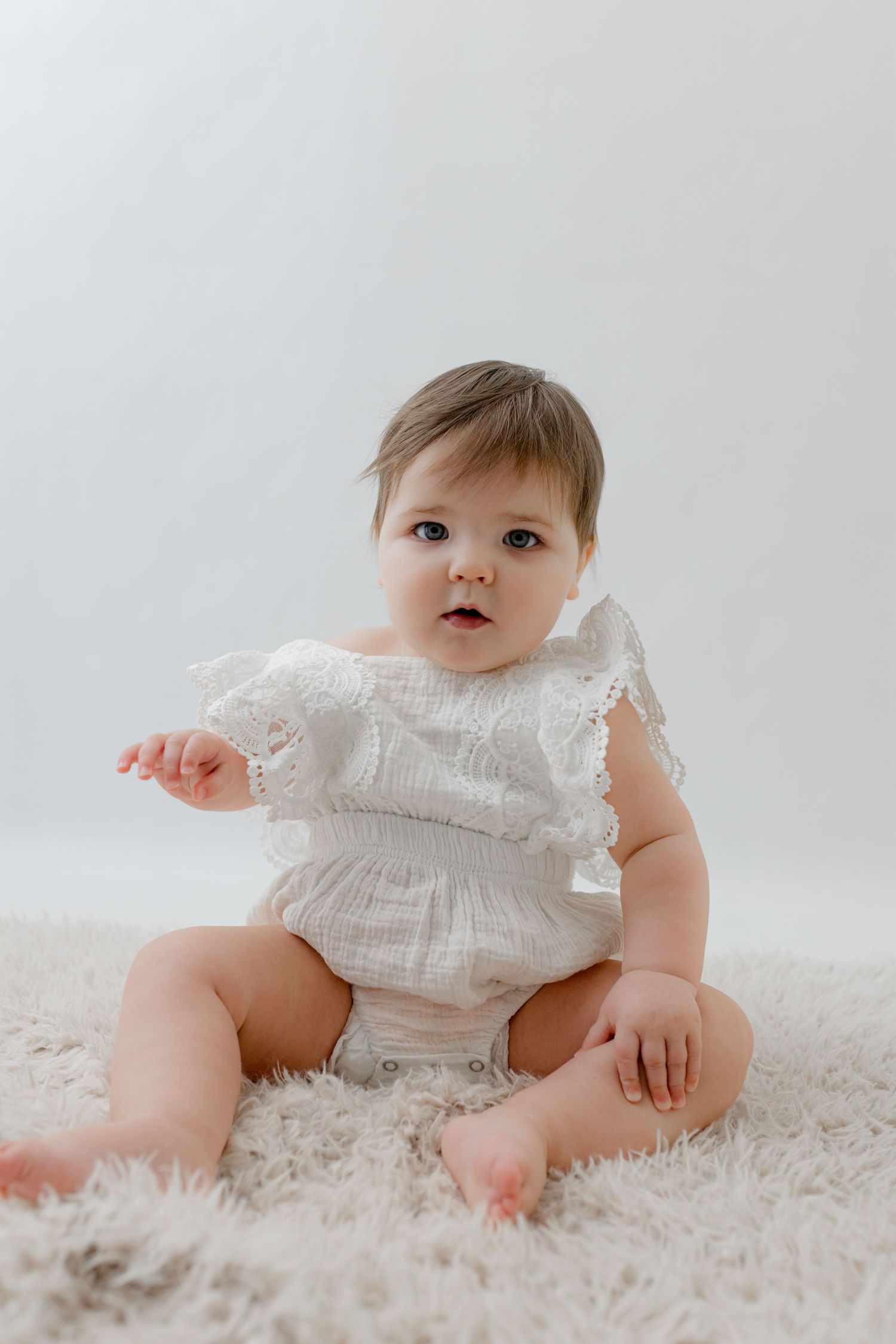 Adorable baby girl in white lace romper sitting on fluffy white rug, looking curiously at camera.