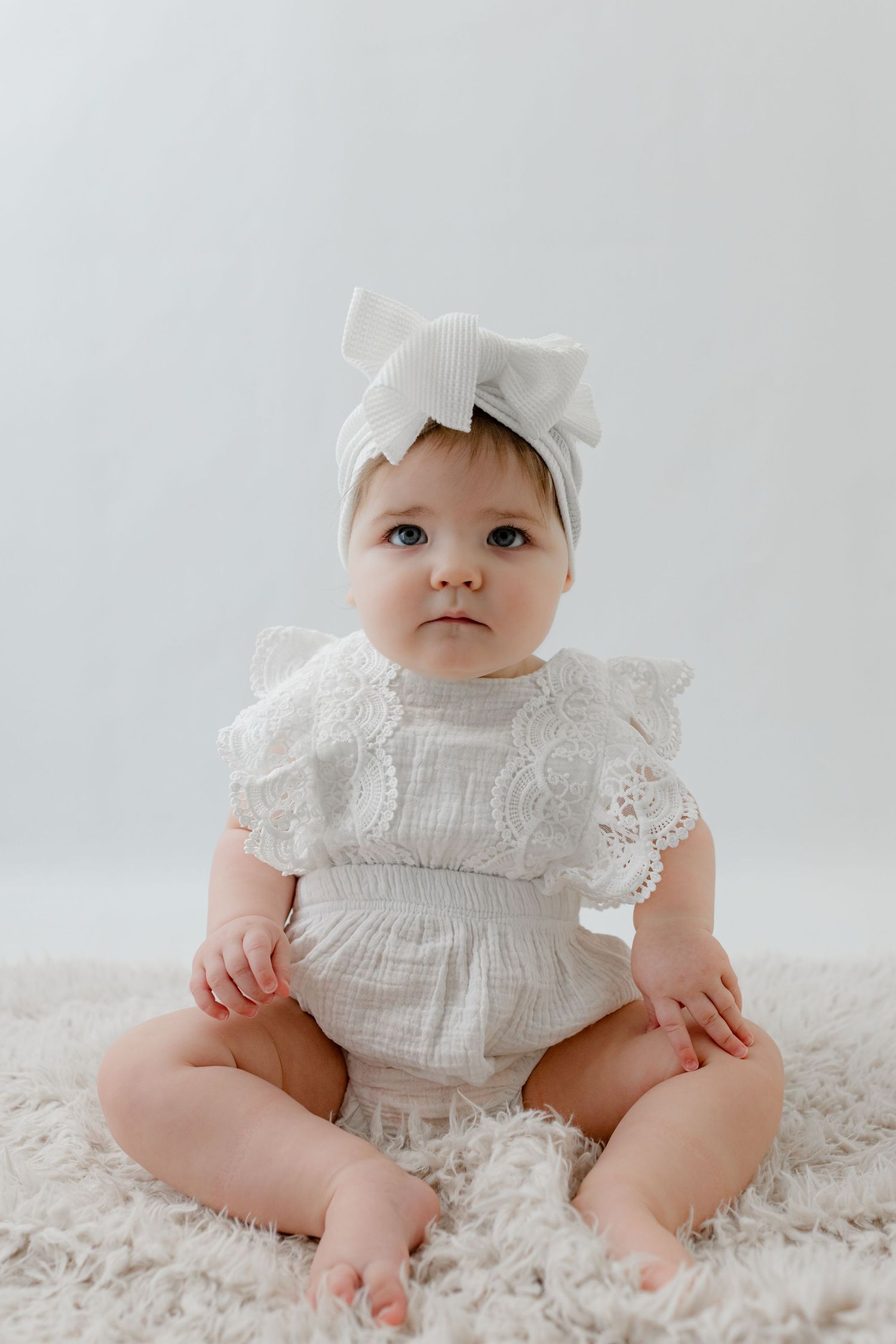 Baby girl in white lace romper and bow headband sits on fluffy rug against light background.