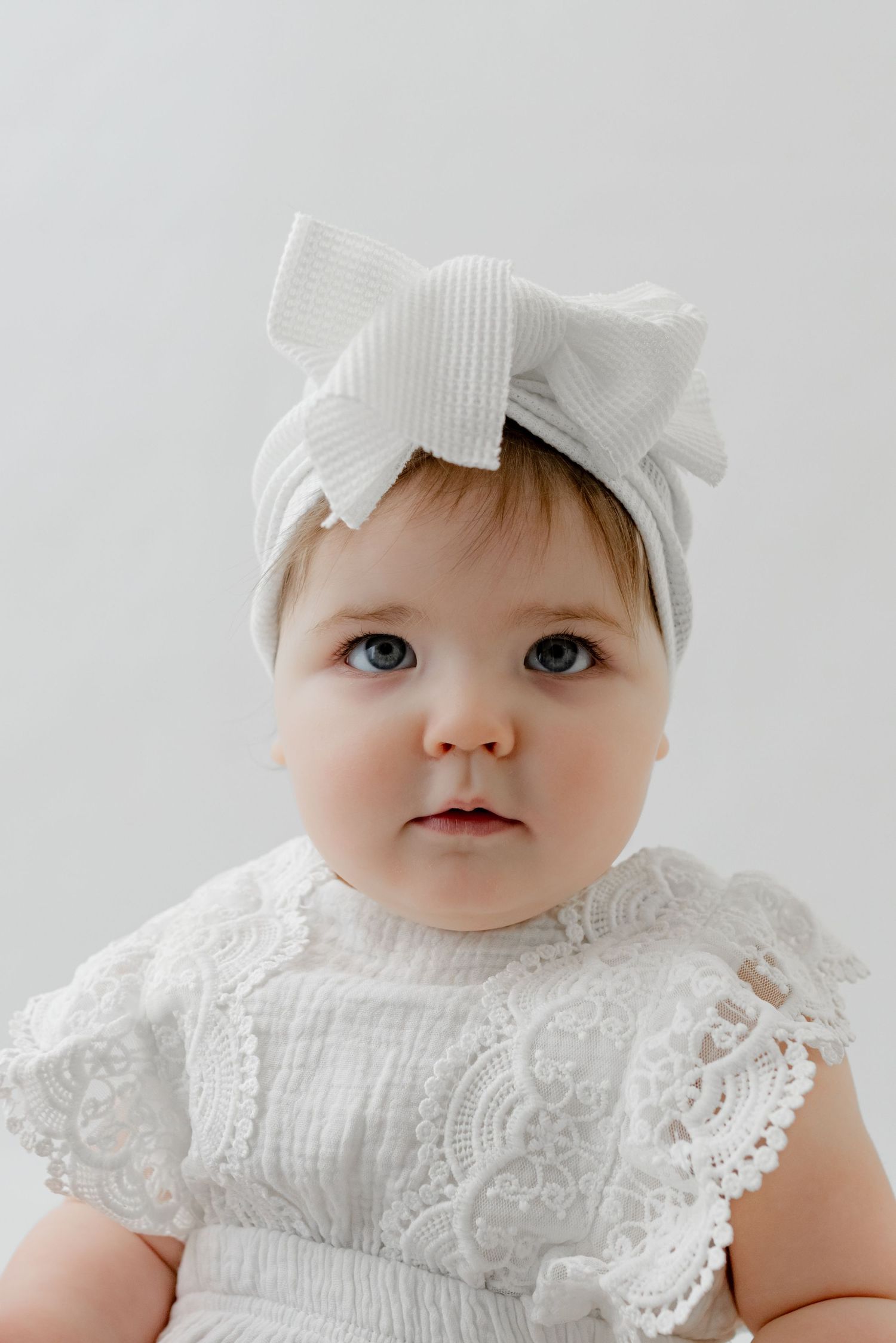 Adorable baby girl in white lace dress and light gray ribbed bow headband, with blue eyes, against white background.