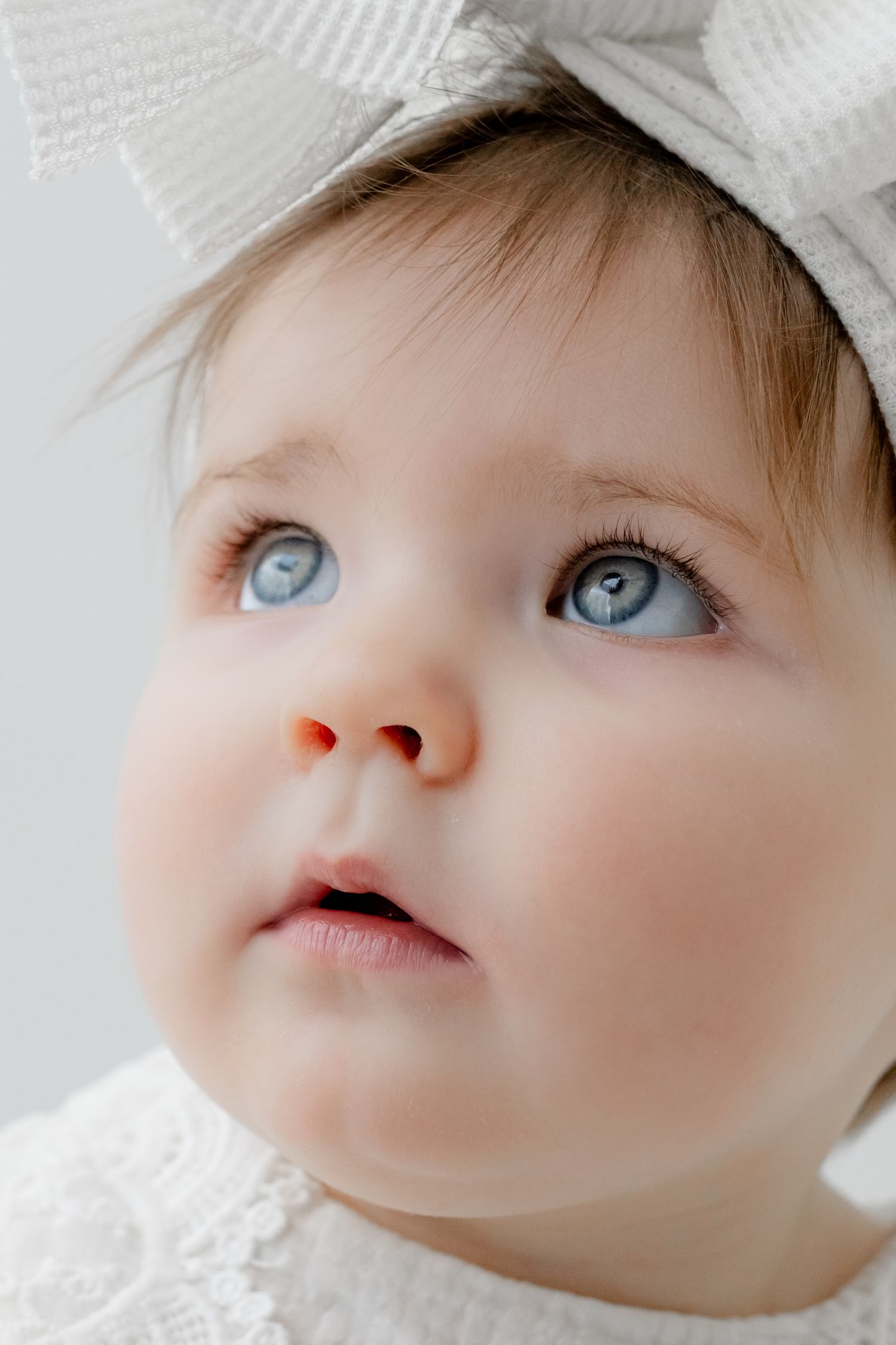 Close-up portrait of a baby girl with blue eyes wearing a white bow headband and white lace dress.