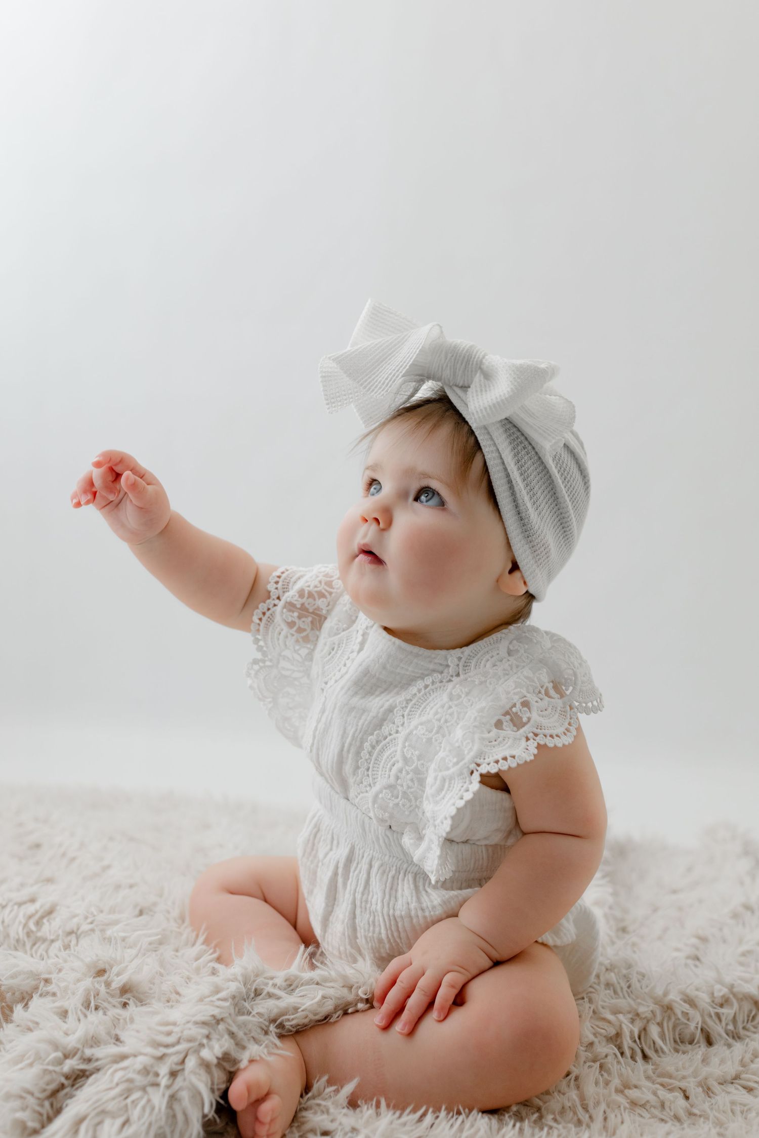 A cute baby girl in a white lace romper and bow headband sits on a fluffy rug, reaching upward.